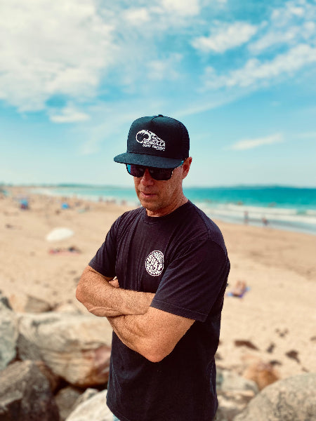 Man wearing a black cap and sunglasses with a beach and ocean in the background
